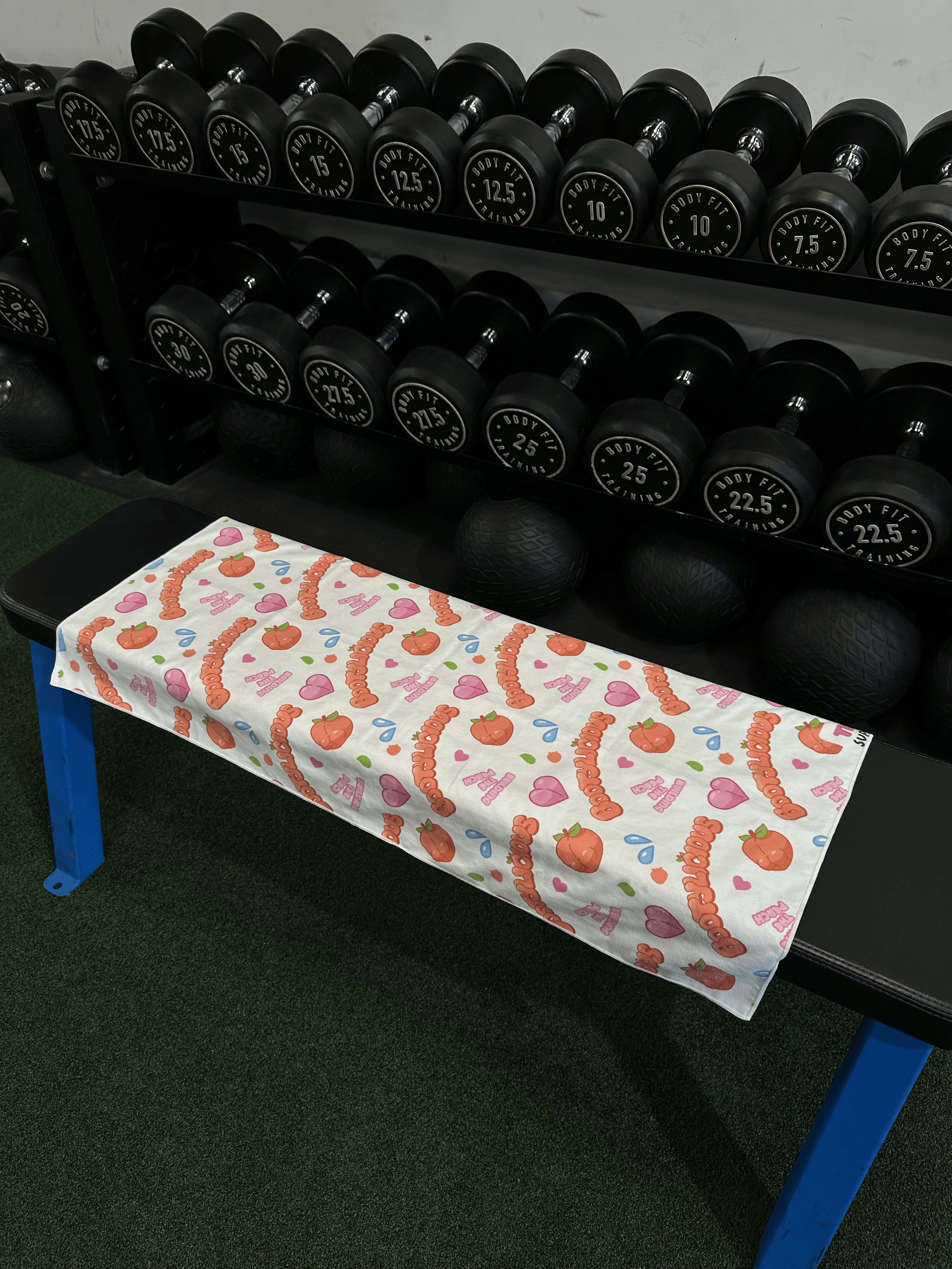 Back of Bootylicious Gym Towel - Repeating Pattern on a bench in front of dumbell rack. 